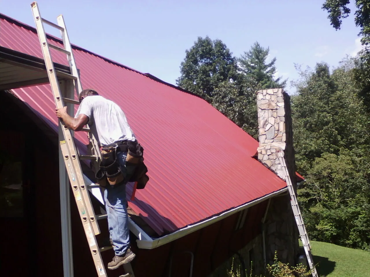 Roofer on a ladder beside a red metal roof and stone chimney — Soffit & Fascia Repair by Roofing Brothers in Moorhead